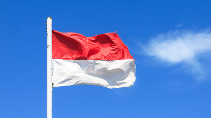 The red and white Indonesia national flag is waving againts clear bright blue sky. little soft white cloud behind.