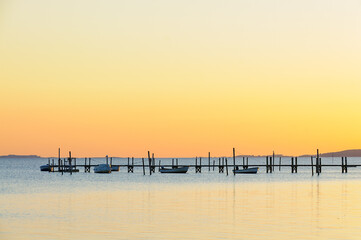 A tranquil shoreline glows orange as the sun sets, casting golden reflections on still waters near wooden piers and boats resting peacefully.