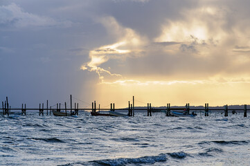 Soft waves gently lap at fishing boats near a stilted dock beneath an enchanting sunset, casting a...