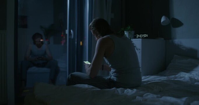 A man in pajama pants uses a mobile phone while sitting on the bed at night. Dark room, home interior