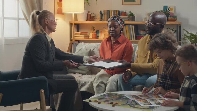 Female social worker giving documents to adoptive Black young parents signing papers and handing them back in living room while two Caucasian little boys leafing through picture book at coffee table