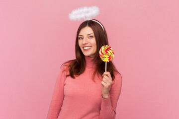 Smiling beautiful happy woman with brown hair and nimb over head, showing colorful lollipop, looking at camera, wearing rose turtleneck. Indoor studio shot isolated on pink background