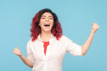 Portrait of extremely happy overjoyed woman with fancy red hair clenched fists, rejoicing, celebrating her victory, success, wearing white shirt. Indoor studio shot isolated on blue background.