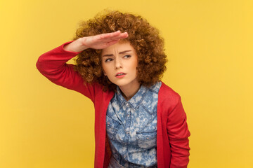 Portrait of serious attractive woman with Afro hairstyle standing with hand at her forehead looking far away with attentive expression. Indoor studio shot isolated on yellow background.