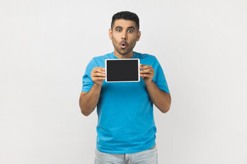 Portrait of amazed surprised unshaven man wearing blue T- shirt standing showing tablet with black bank screen, advertisement area. Indoor studio shot isolated on gray background.