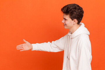 Portrait of young man in white hoodie giving hand for handshake as sign of greeting, smiling sincerely, looking very glad to see person he meeting. Indoor studio shot isolated on orange background.