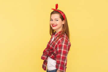 Side view portrait of joyful woman with red lips ,standing looking at camera, expressing happiness, wearing checkered shirt and headband. Indoor studio shot isolated on yellow background.