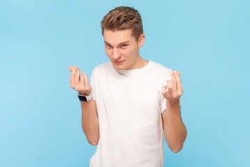 Portrait of cunning handsome man wearing white t-shirt standing showing money gesture, asking his salary, looking at camera with sly look. Indoor studio shot isolated on blue background