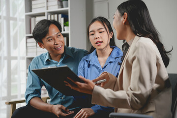 Happy asian couple discussing terms of contract with professional realtor while sitting on sofa in modern office