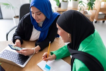 Two women in hijabs effectively planning their goals in a modern office environment, using laptops, notes, and a goal-setting approach.