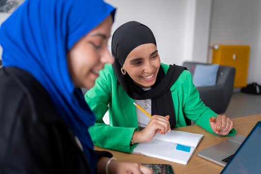 Two women in hijabs smiling and working together in a modern office environment, using laptops and taking notes for their project.