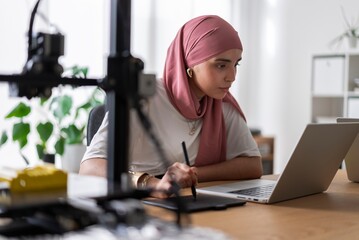 A woman in a hijab is intently using a laptop and graphic tablet in a well-lit office environment, concentrating on her work and digital creation
