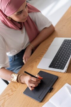 A woman wearing a hijab works on a graphic tablet while using a laptop She is focused and appears to be creating digital art or designing in a modern workspace