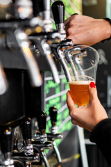 A close-up of a hand with red nail polish pouring beer from a tap into a glass at a bar. The focus is on the pouring action, with beer taps and the glass prominently visible.