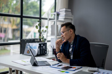 A man in a blue jacket is sitting at a desk with a laptop and a tablet