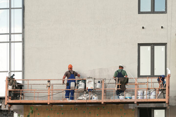 Facade plastering of a multi-storey building. Workers from a construction cradle at height apply...