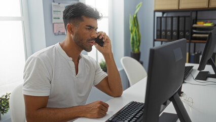 Young hispanic man talking on phone while working at computer in modern office workspace