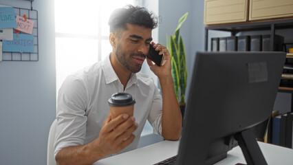 Fototapeta premium Young man with coffee talking on mobile phone at desk in modern office, working on computer, handsome adult in professional workplace.