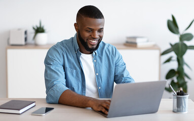 Distance Job. Millennial African American Man Working Online On Computer At Home Office, Handsome Smiling Black Freelancer Guy Sitting At Desk And Looking At Laptop Screen, Free Space