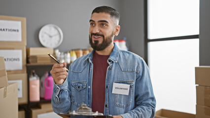 A bearded man volunteers at an indoor workplace, holding a clipboard in a denim jacket labeled...