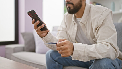 A handsome man with a beard using a smartphone and holding a credit card indoors, suggesting online shopping at home.