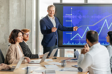 A businessman stands in front of a large screen displaying a graph with an upward trend, pointing to the data. He is presenting to a group of colleagues seated at a conference table.