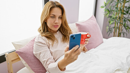 Hispanic woman sitting in her bedroom drinking coffee and using smartphone