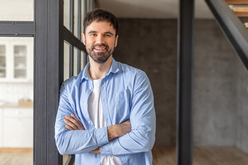 A man stands in a modern building, leaning against a glass wall with his arms crossed. He is wearing a light blue button-down shirt, a white t-shirt, and he is smiling warmly, copy space