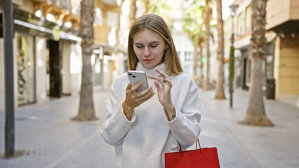 A young caucasian woman with blonde hair and blue eyes using a smartphone on a sunny urban street while shopping.