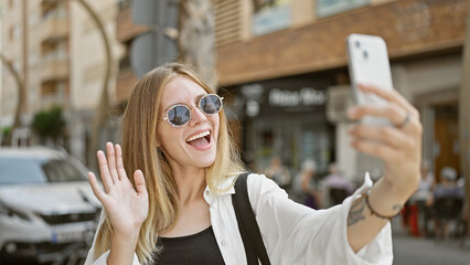 A cheerful blonde woman in sunglasses taking a selfie on a busy city street.