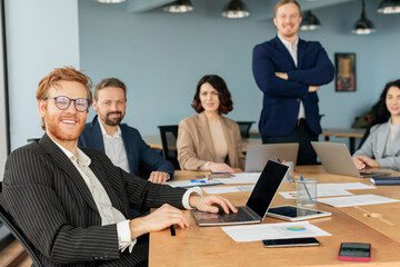 A team of business professionals is gathered around a large conference table, collaborating and working on projects. One man, with a red beard and glasses, is smiling and working on a laptop