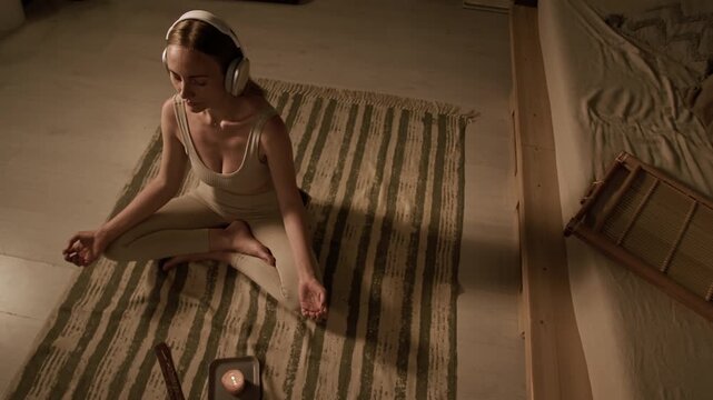 Top rotating view of Caucasian woman in beige activewear meditating at home on floor, sitting in sukhasana pose and listening to relaxing music on headphones, decorated room at dawn with soft light