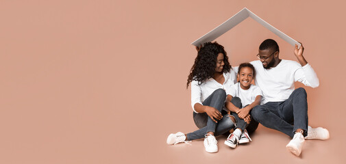 Adoption concept. African american man and woman, adoptive parents holding symbolic roof above little girl, sitting together on yellow background. © Prostock-studio