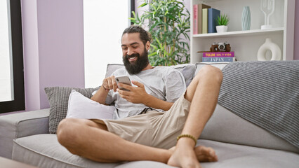 Handsome bearded hispanic man relaxing and smiling while using his smartphone on a comfy sofa in a modern living room.