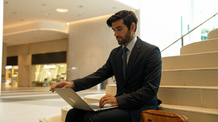 Skilled investor working or planing strategy by using laptop at stair. Professional business man wearing suit while working and typing data analysis by using laptop at modern hotel. Exultant.