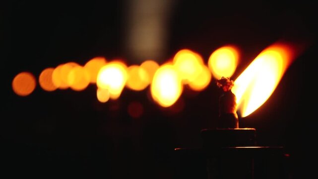A close up shot of a line of lit torches, their flames glowing warmly in the dark night. The foreground torch is in sharp focus, while the others blur into the background, creating a serene
