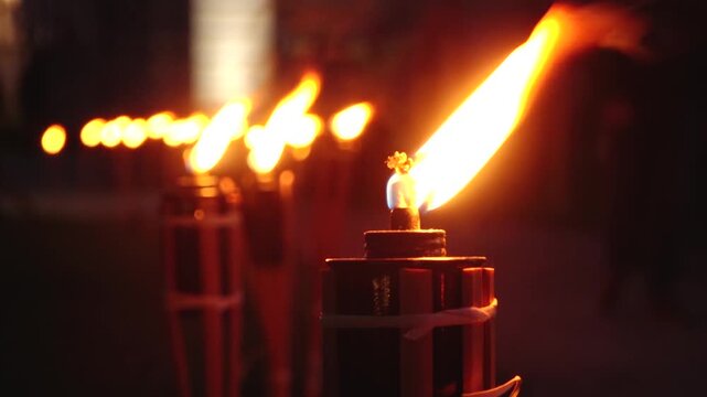 A close up shot of a line of lit torches, their flames glowing warmly in the dark night. The foreground torch is in sharp focus, while the others blur into the background, creating a serene