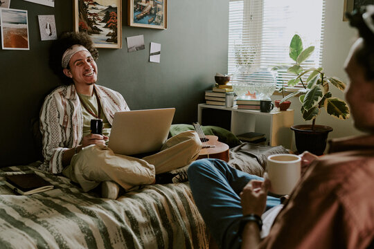 Curly-haired student sitting with laptop on bed and loathing out loud while his roomie telling joke and drinking coffee