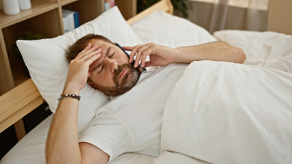 A bearded mature man feeling unwell while talking on the phone in a bedroom setting.