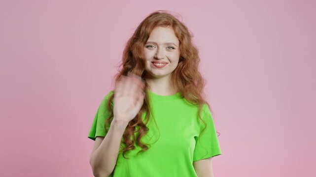 Friendly young woman waving hand - hello, or salute, chao, adios. Parting, say bye to camera. Beautiful summer lady on pink studio background