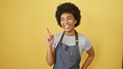 Smiling african american woman with curly hair posing in denim apron against a yellow background.