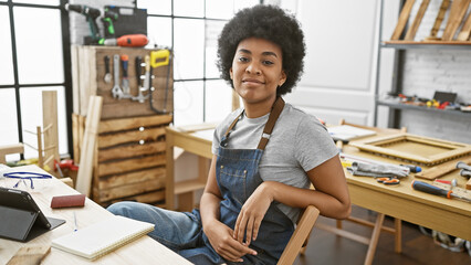 Confident female carpenter with curly hair posing in a well-organized workshop full of woodworking...