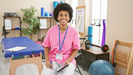 A smiling african american woman healthcare professional seated in a rehab clinic room, displaying confidence and approachability.