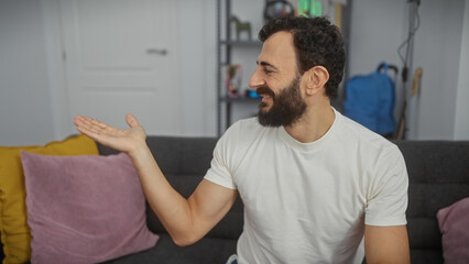 A bearded man in a white shirt smiles and gestures with an outstretched hand in a modern living room setting