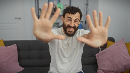 Naklejka premium Bearded man gesturing stop with hands in a modern living room, looking playful and smiling.