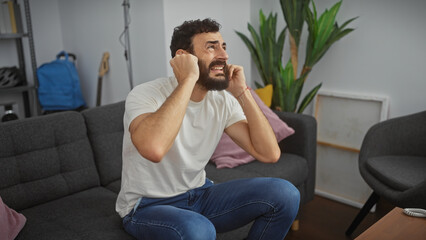 A distressed man covers his ears while sitting on a gray couch indoors, displaying discomfort from loud noise.