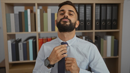 Handsome young hispanic man with a beard adjusting his tie in an office building with bookshelves...