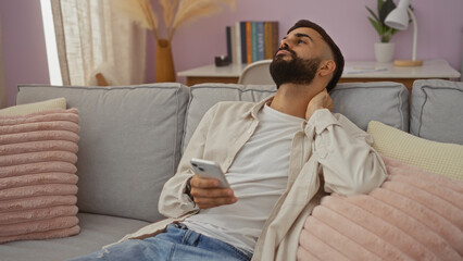 Young man with a beard sitting in a cozy living room, touching his neck in discomfort, while...