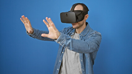 Fototapeta premium Hispanic man with beard using virtual reality headset against blue background, exploring vr technology wearing casual denim attire.