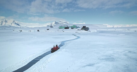 Scientist from polar station base riding zodiac boat in melted steam. Frozen ocean ice, mountains and science buildings in background. South Pole tourism, travel, expedition. Aerial drone panorama